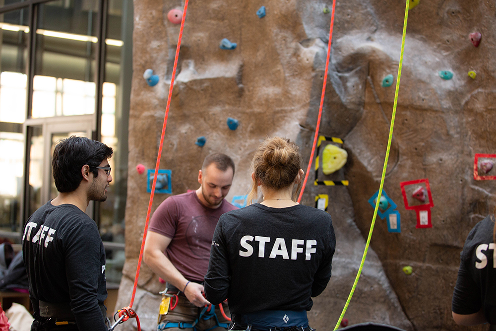 Two individuals with Staff written on their shirts helping another individual prepare to climb the climbing wall at the Campus Recreation and Wellness Center