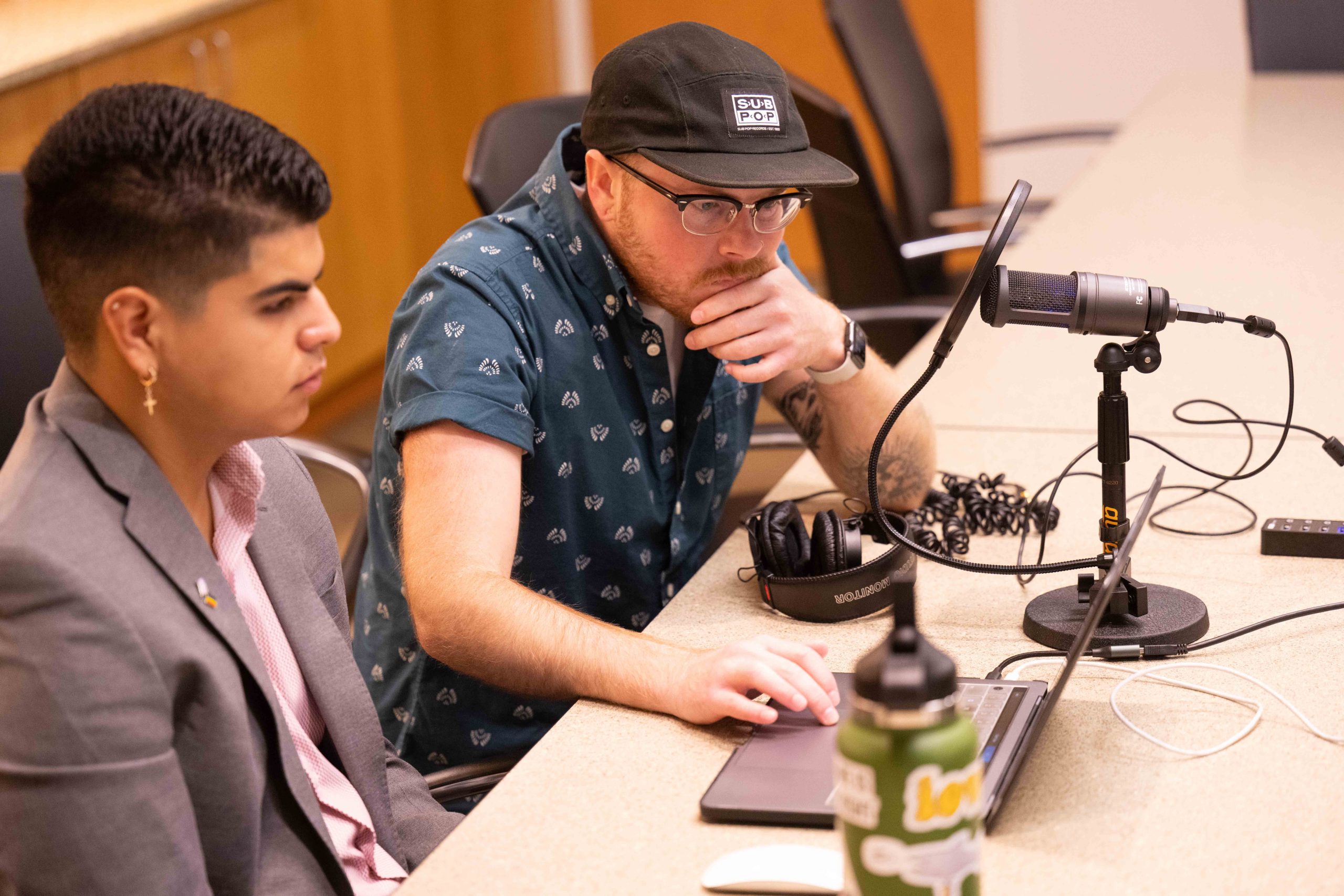 Two people sitting in front of a microphone, looking at a laptop computer.