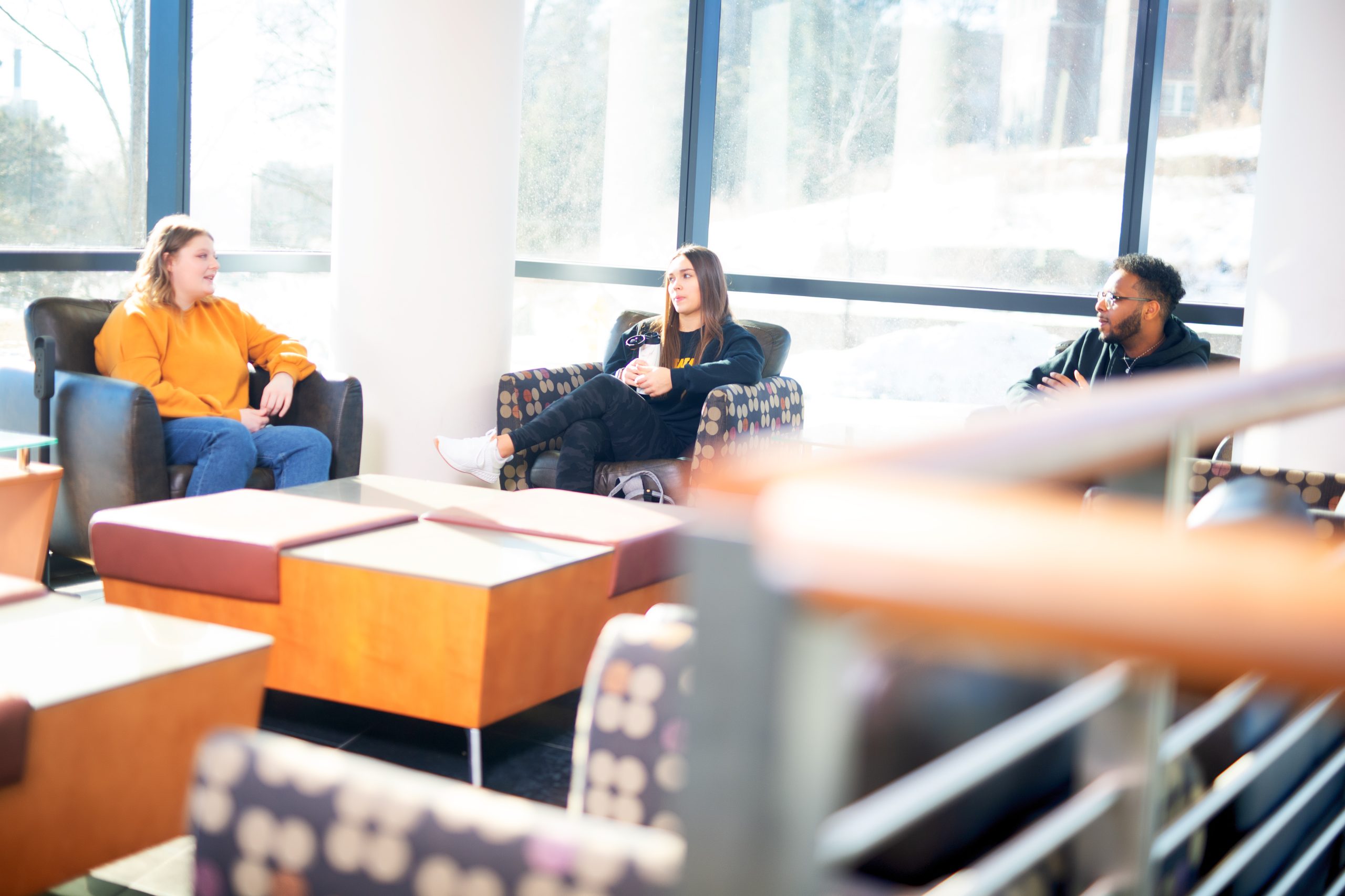 Three students sitting on chairs in a bright study area, talking to each other.