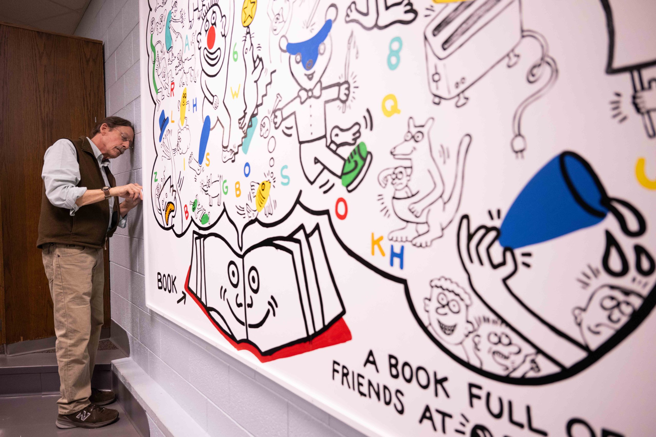 A person is attaching a large printed and framed picture onto a wall. The pictures contains colorful, drawn figures on white background.