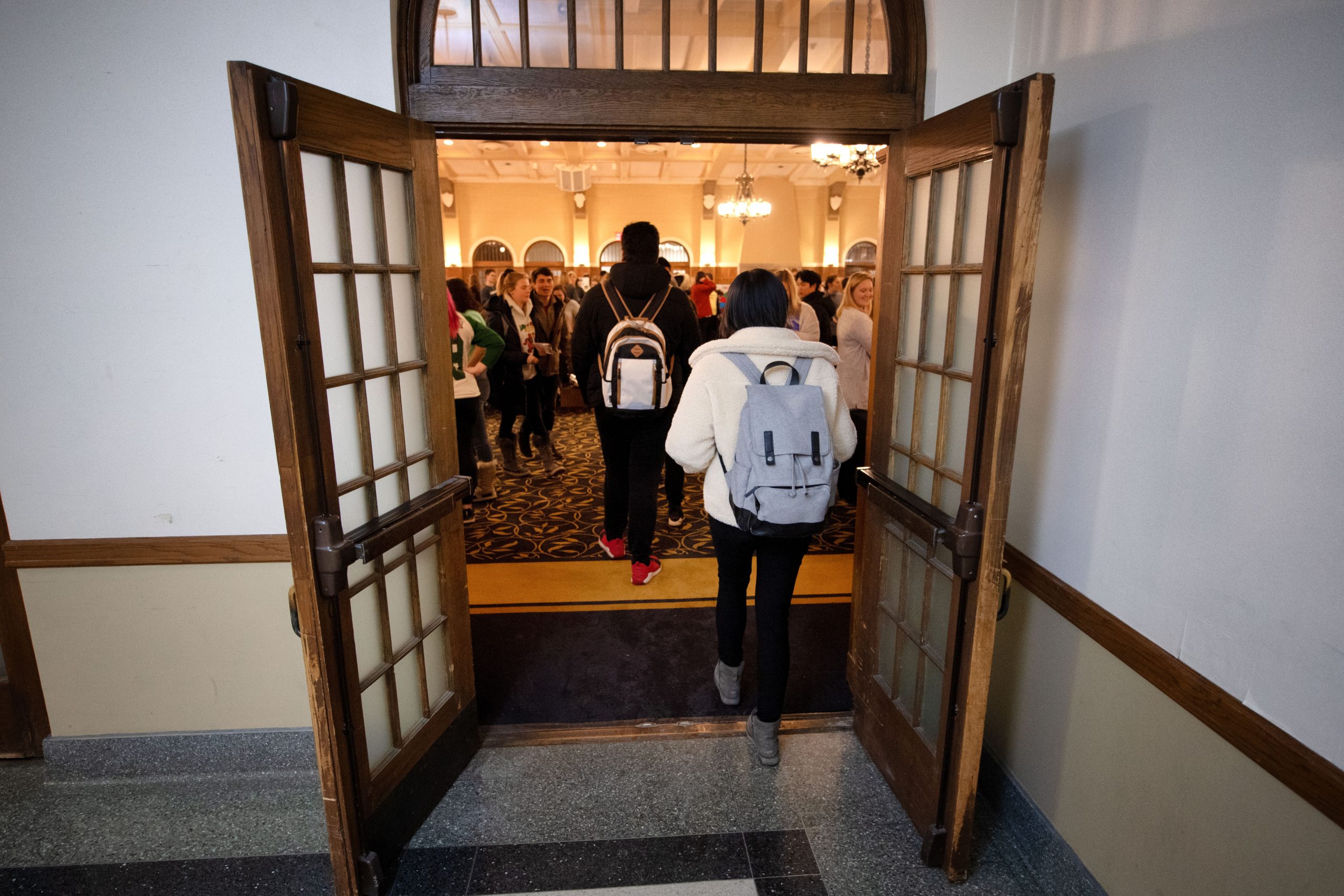 A group of students are entering a large banquet hall. They are carrying backpacks.