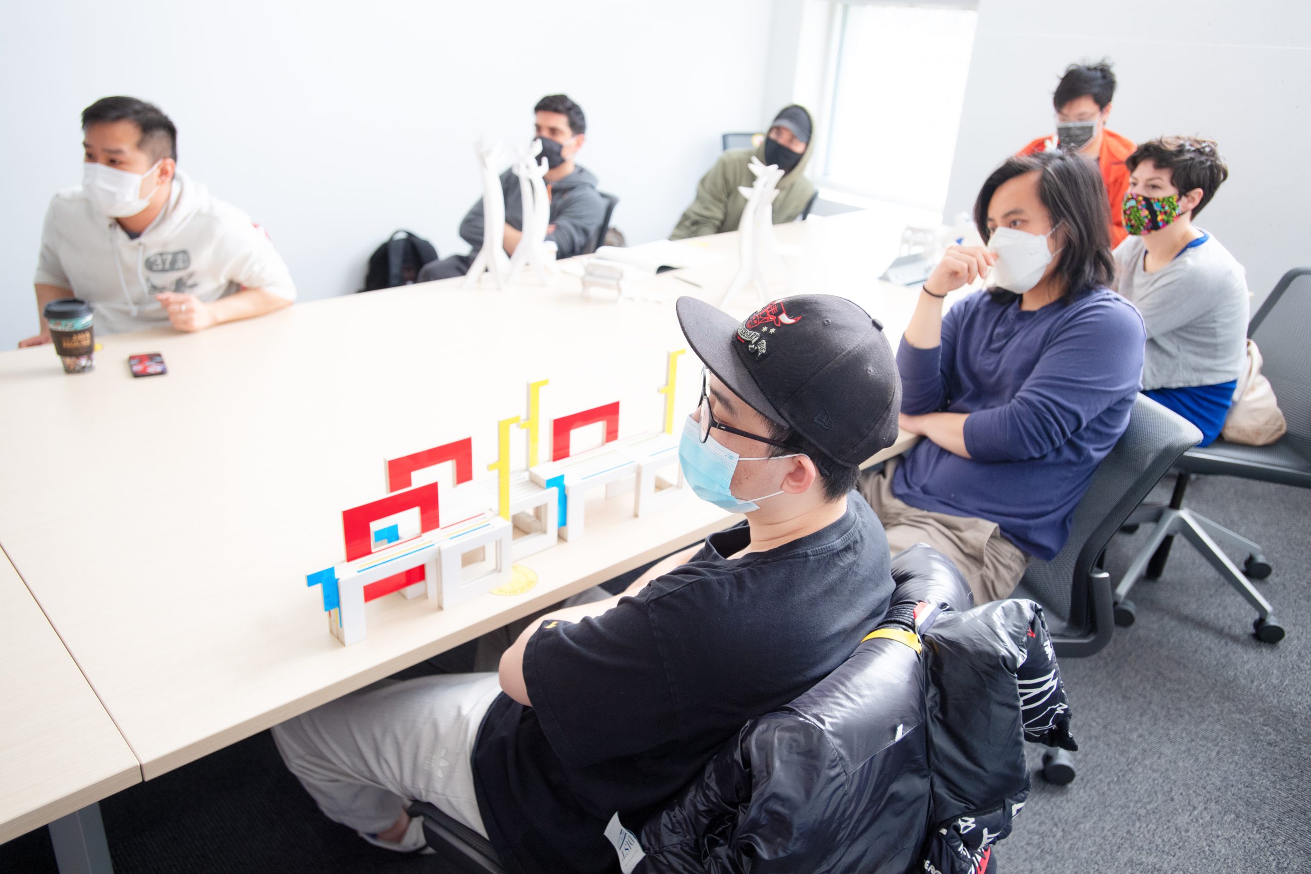 Seven students, wearing masks are sitting around a long table. They are looking towards something outside the frame.