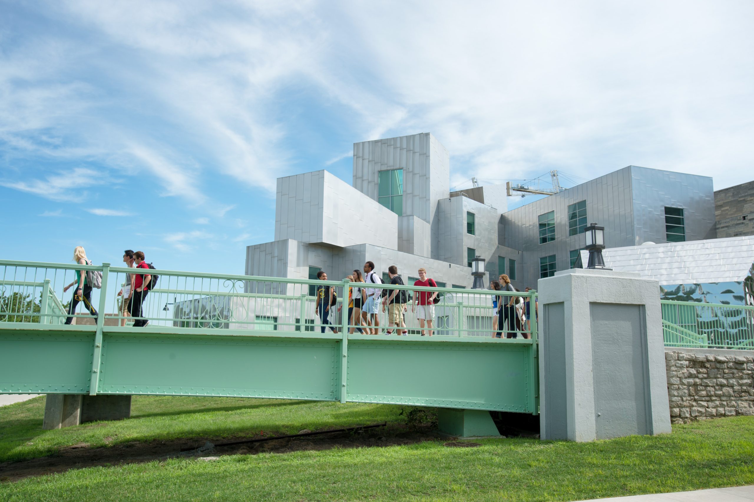 A group of young people crossing a green bridge over the Iowa River in downtown Iowa City. In the background is blue sky and a cluster of silver buildings.