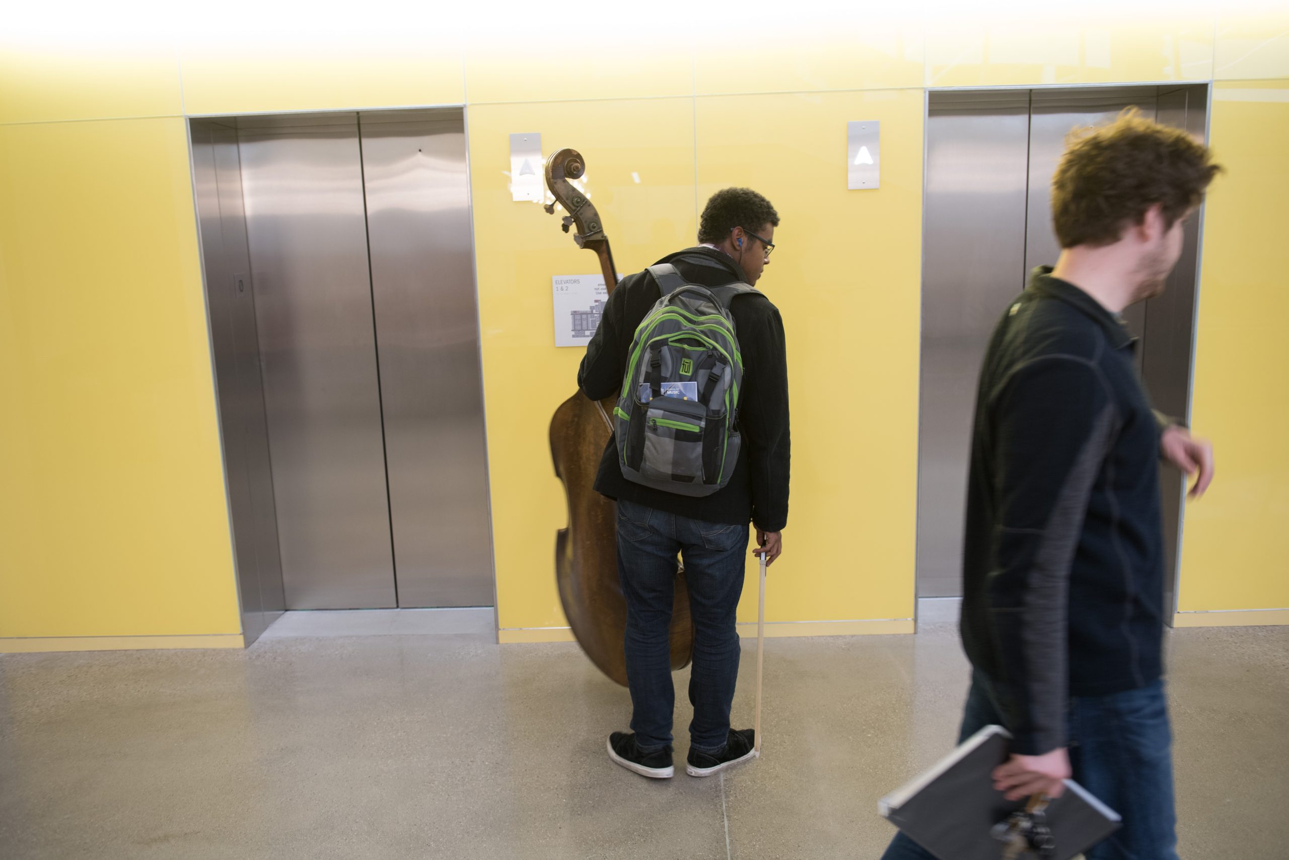 A student, holding a double bass musical instrument while waiting for an elevator. Another student, holding a book is walking past.