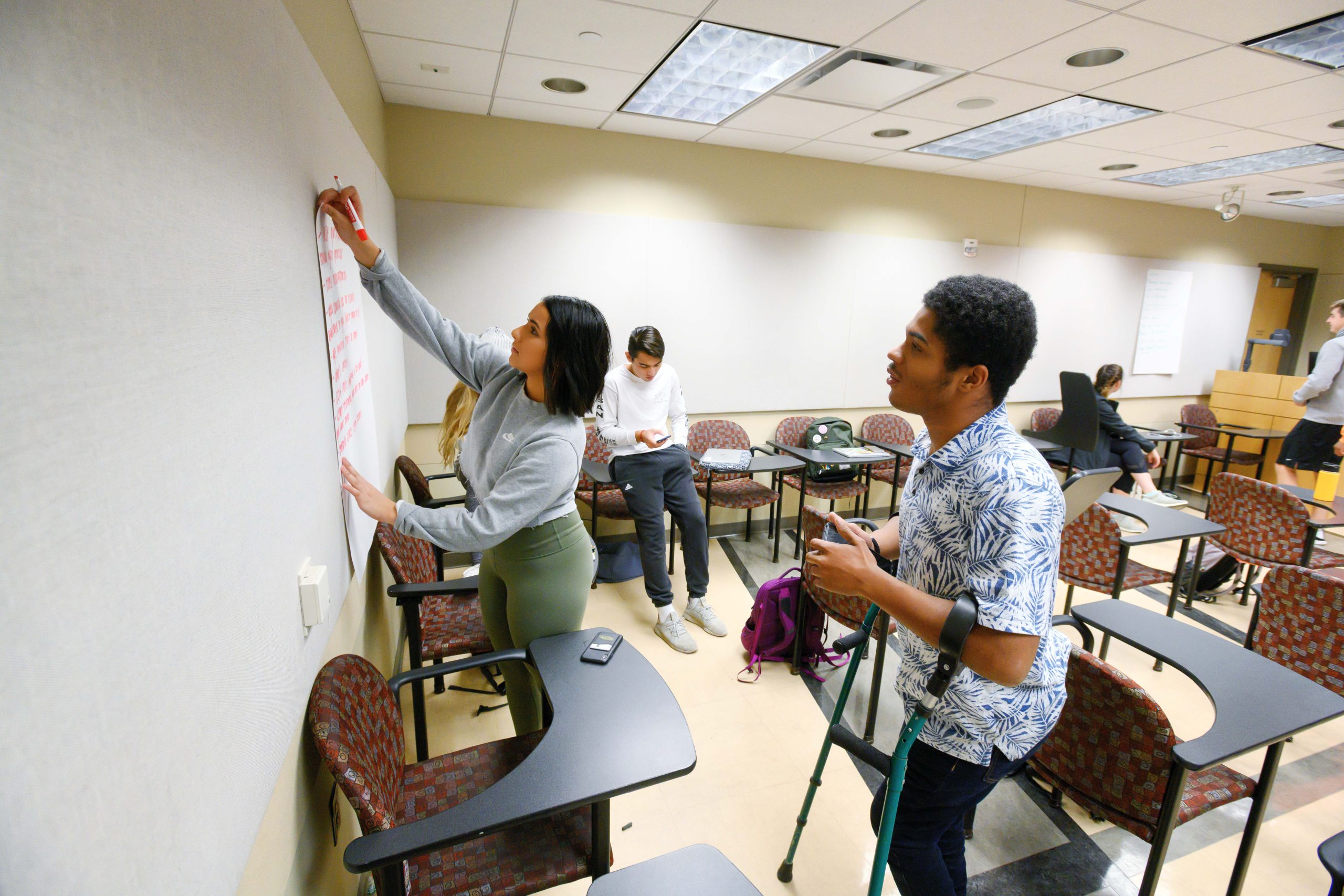 A group of three students attaching a large sheet of paper to a white wall. The paper contains handwritten notes in red ink. Two more students are in the background, talking to each other.