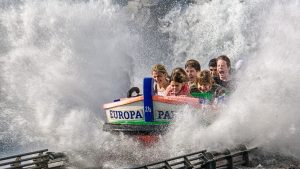 A group of people on a water ride at Europa Park, splashing through a large wave. Their faces show excitement and joy as water sprays all around them. The boat is brightly colored with blue and green details, creating a thrilling amusement park moment