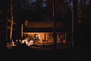 A cozy wooden cabin in the woods at night, softly illuminated by interior lights and a nearby campfire. A group of people is gathered around the fire, sitting in chairs and enjoying the warmth. The surrounding trees and darkness create a peaceful, rustic atmosphere.