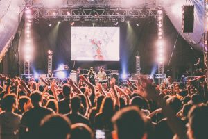 A lively concert scene with a band performing on stage under bright lights. The crowd is raising their hands, cheering, and dancing. A large screen behind the performers displays visuals, enhancing the energetic atmosphere of the live music event.