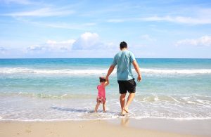 A father and his young daughter walk hand in hand along the shoreline of a sandy beach, stepping into the gentle waves. The sky is bright blue with scattered clouds, and the ocean stretches out to the horizon.