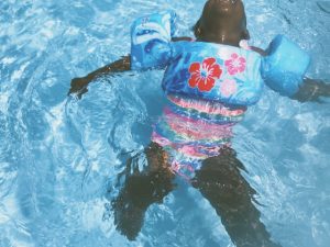 A young child floating on their back in a swimming pool, wearing a colorful floral swimsuit and blue arm floaties. The child appears relaxed, with arms outstretched and head tilted back, enjoying the water.