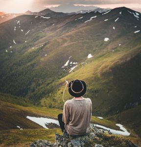 A person wearing a black hat and a beige sweater sits on a rocky ledge, gazing at a vast mountain landscape. The rolling green hills and snow-capped peaks stretch into the distance under a dramatic sky, with rays of sunlight breaking through the clouds.