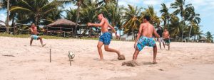 Four young men playing beach soccer on a sunny day, wearing colorful swim trunks. The sandy field is surrounded by palm trees, with huts and a fence in the background. The sky is bright blue with scattered clouds