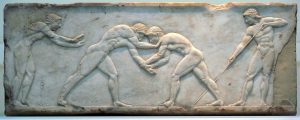 a marble relief depicting ancient Greek athletes engaged in wrestling. The artwork shows two central figures locked in a grappling stance, demonstrating their muscular physiques and physical struggle. To the left, a third figure appears to be a trainer or fellow athlete, extending his arms, possibly offering guidance or encouragement. On the right, a fourth figure, likely a referee or judge, holds a staff, overseeing the competition. The relief showcases the Greek admiration for athleticism and competition, often associated with events like the Olympic Games. The intricate carving and details in the figures' hair, musculature, and poses reflect classical Greek artistic conventions.