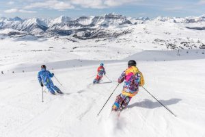 Three skiers wearing colorful outfits carving through fresh snow on a mountainside, with a stunning backdrop of snow-covered peaks under a bright blue sky. The scene captures the thrill of skiing in a breathtaking alpine landscape.
