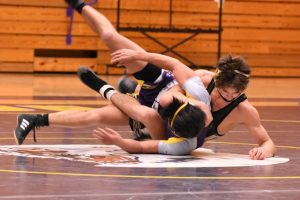 Two high school wrestlers engaged in a match on a gymnasium mat. One wrestler in a black singlet is pinning the other, who is wearing a purple and yellow singlet, while executing a takedown. The gym features a wooden backdrop with team banners.