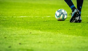 A close-up of a soccer player's legs and feet on a green grass field, controlling a white and green soccer ball. The player is wearing dark athletic pants and black cleats.