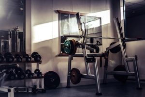 A gym with weightlifting equipment, including a squat rack, barbells, and dumbbells, illuminated by natural light from a nearby window.
