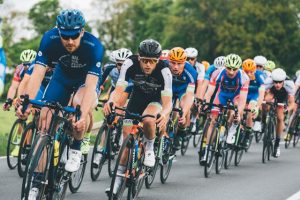 A group of professional cyclists in colorful jerseys and helmets racing on a paved road surrounded by greenery. The cyclists in the foreground are intensely focused, gripping their handlebars as they ride closely together in a competitive formation.