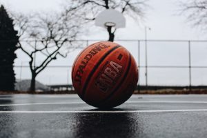 A Spalding NBA basketball resting on a wet outdoor court with a basketball hoop in the background. The overcast sky and bare trees create a moody atmosphere, suggesting a cold or rainy day