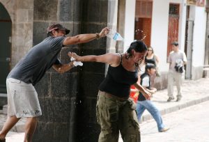 A man playfully pours water from a plastic bottle over a laughing woman in a public square.