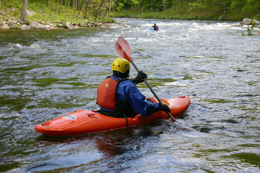 A man with oar in his hand is kayaking downstream in a shallow fast-flowing river.