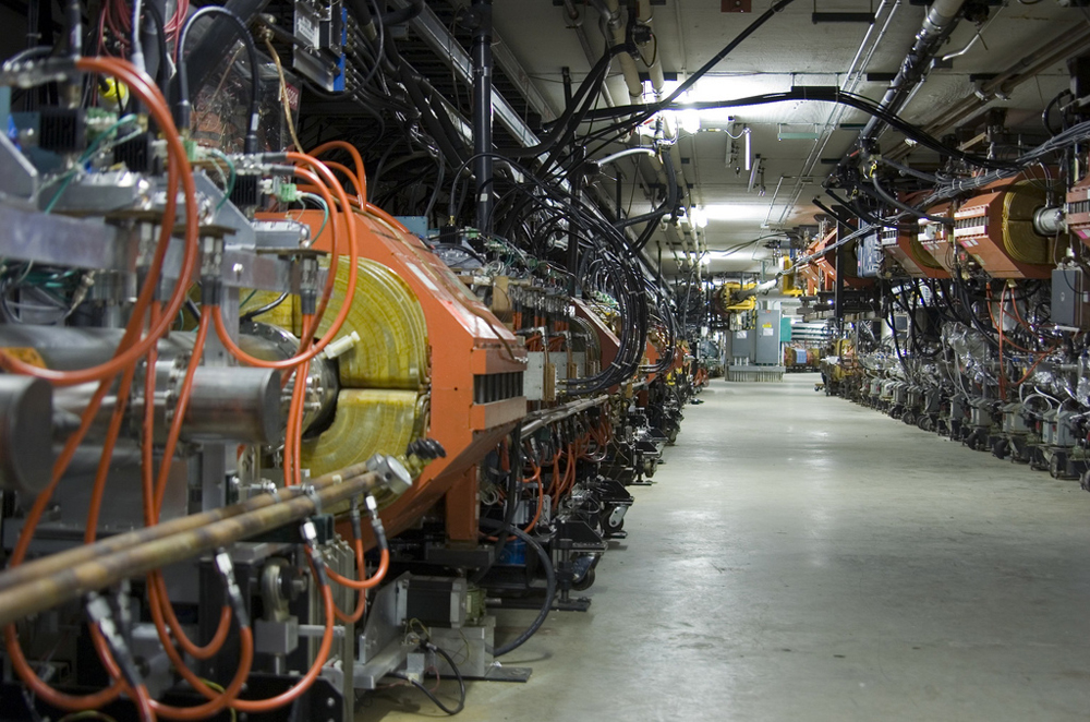 A view of a section of the accelerator at Fermilab. Down each side of a long corridor are tubes surrounded by orange magnets. Lots of tubes and wires and other electronics are visible.