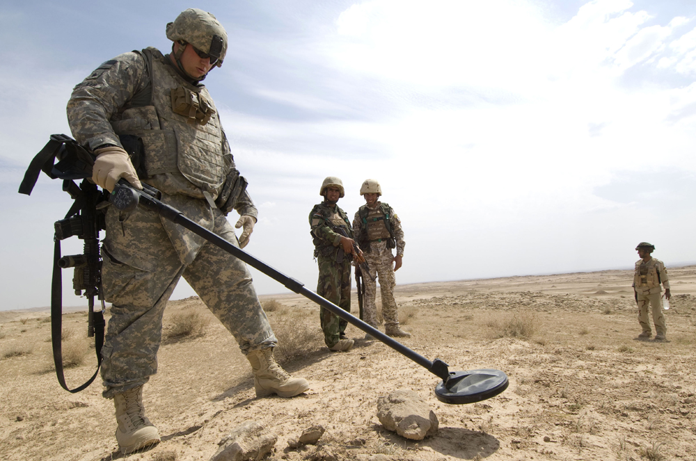 ]Photograph of several soldiers in an open field. One soldier is searching for explosives by scanning the surface using a metal detector.