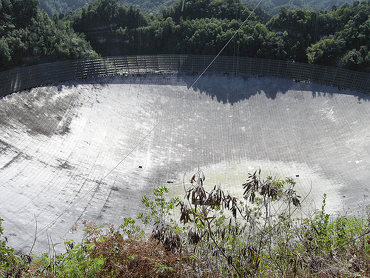 The figure shows a photograph from above looking into the Arecibo Telescope in Puerto Rico. It is a huge bowl-shaped structure lined with reflecting material. The diameter of the bowl is three times as long as a football field. Trees can be seen around the bowl, but they do not shade the bowl significantly.
