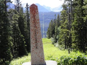 A forest is divided in two by a natural space. A stone border marker appears in the front and a mountain range in the back
