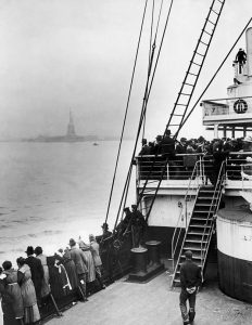 A group of immigrants are in a ship at sea and the Statue of Liberty can be seen in the background